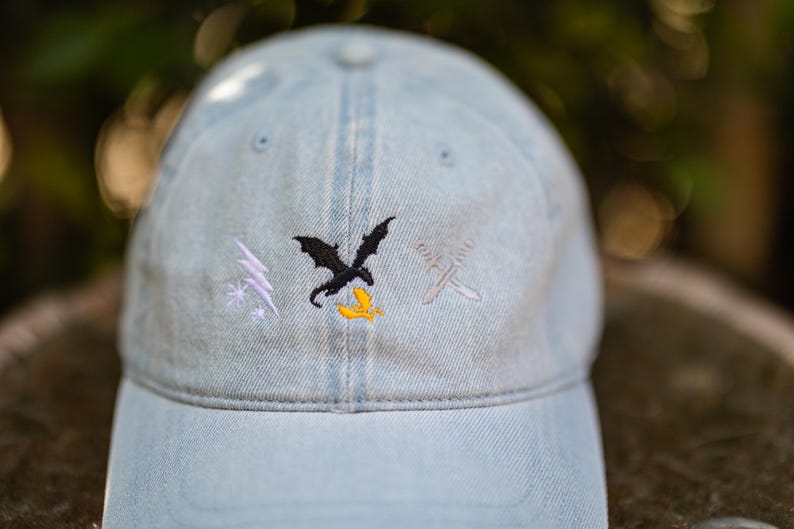 May include: A light blue denim baseball cap featuring three embroidered designs on the front panel: a white lightning bolt with a star, a black dragon silhouette above a small yellow flame, and two crossed silver swords.