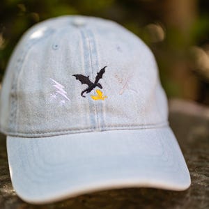 May include: A light blue denim baseball cap featuring an embroidered design on the front. The embroidery includes a black dragon, a white lightning bolt with small stars, a yellow flame, and a pair of crossed white swords on the right side.