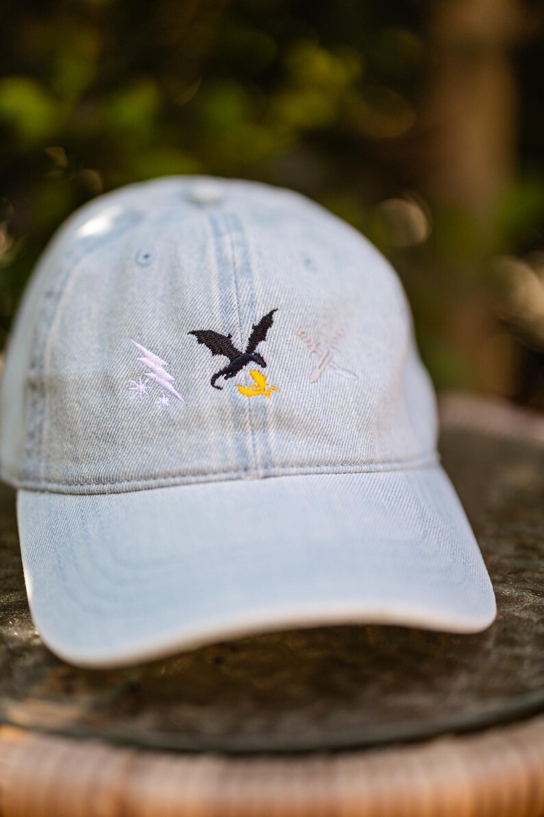 May include: A light blue denim baseball cap featuring an embroidered design on the front. The embroidery includes a black dragon, a white lightning bolt with small stars, a yellow flame, and a pair of crossed white swords on the right side.
