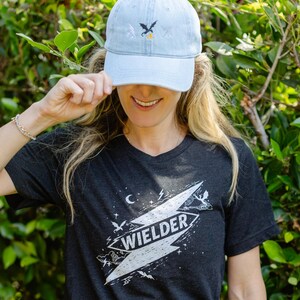 May include: A light blue denim baseball cap with a small embroidered dragon and lightning bolt design, paired with a dark charcoal gray t-shirt featuring a large white graphic of a lightning bolt containing the text WIELDER and small dragons.
