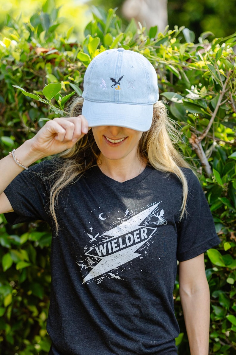 May include: A light blue denim baseball cap with a small embroidered dragon and lightning bolt design, paired with a dark charcoal gray t-shirt featuring a large white graphic of a lightning bolt containing the text WIELDER and small dragons.