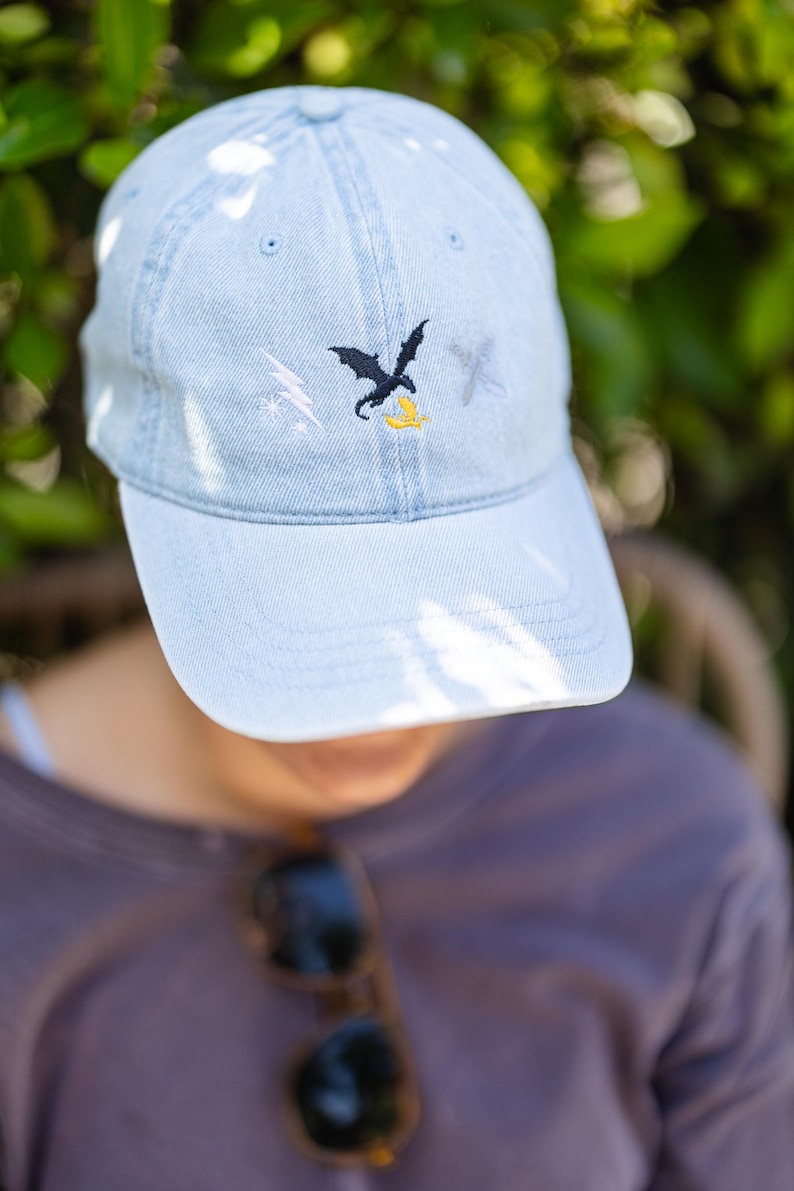 May include: A light blue denim baseball cap featuring an embroidered design on the front. The embroidery includes a white lightning bolt, a black dragon silhouette, and a small yellow banana shape, with a faint grey cross symbol to the right.