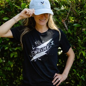 May include: A light blue baseball cap with an embroidered dragon and moon design, paired with a black short-sleeve t-shirt featuring a white graphic print of a lightning bolt, stars, and the word WIELDER centered on the chest area.