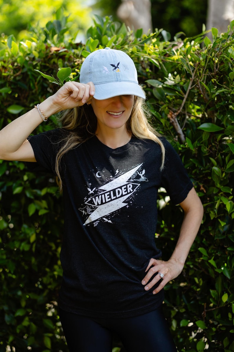 May include: A light blue baseball cap with an embroidered dragon and moon design, paired with a black short-sleeve t-shirt featuring a white graphic print of a lightning bolt, stars, and the word WIELDER centered on the chest area.