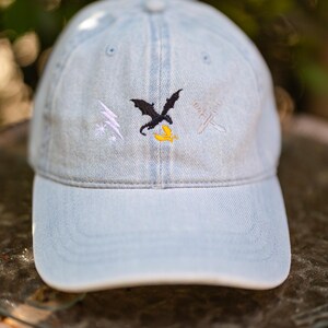 May include: A light blue denim baseball cap featuring an embroidered design on the front. The embroidery includes a white lightning bolt, a black dragon silhouette above a yellow bird, and two crossed silver swords on the right side.