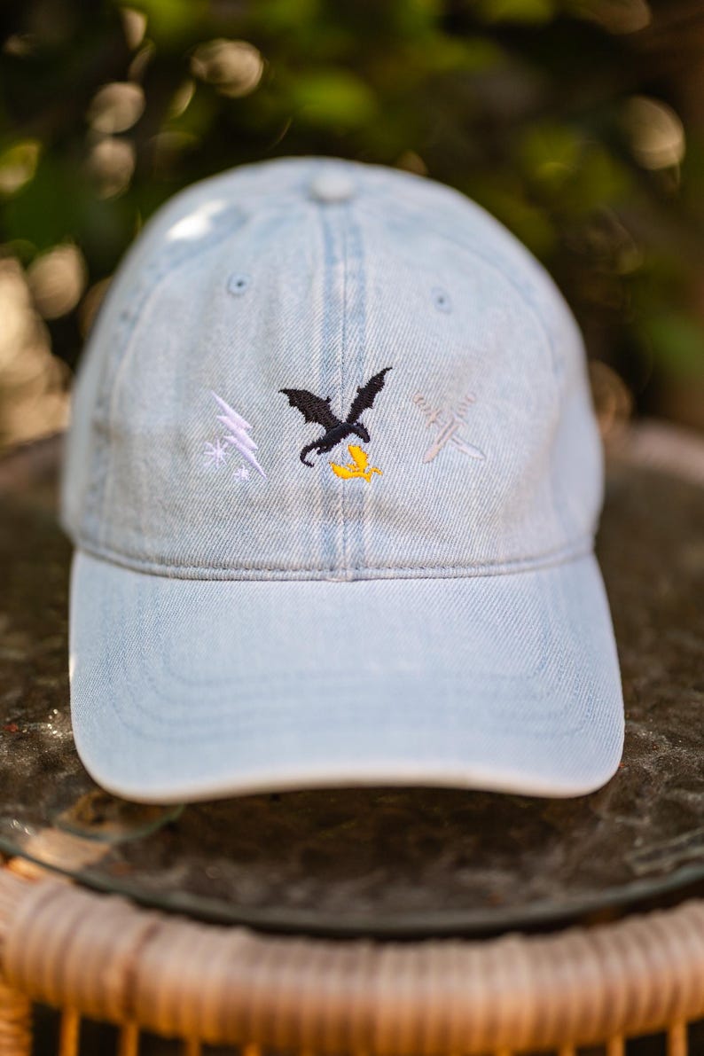 May include: A light blue denim baseball cap featuring an embroidered design on the front. The embroidery includes a white lightning bolt, a black dragon silhouette above a yellow bird, and two crossed silver swords on the right side.