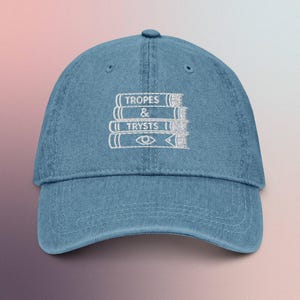 May include: A light blue denim baseball cap with a curved peak. The cap has white embroidered text and a graphic of stacked books with the words "TROPES & TRYSTS". The cap has a classic six-panel design with a button on top.