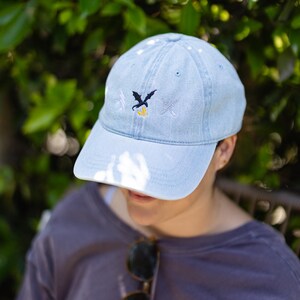 May include: A light blue denim baseball cap featuring an embroidered design on the front. The embroidery consists of a small black dragon silhouette above a yellow flame, flanked by a white lightning bolt on the left and a white sword on the right.