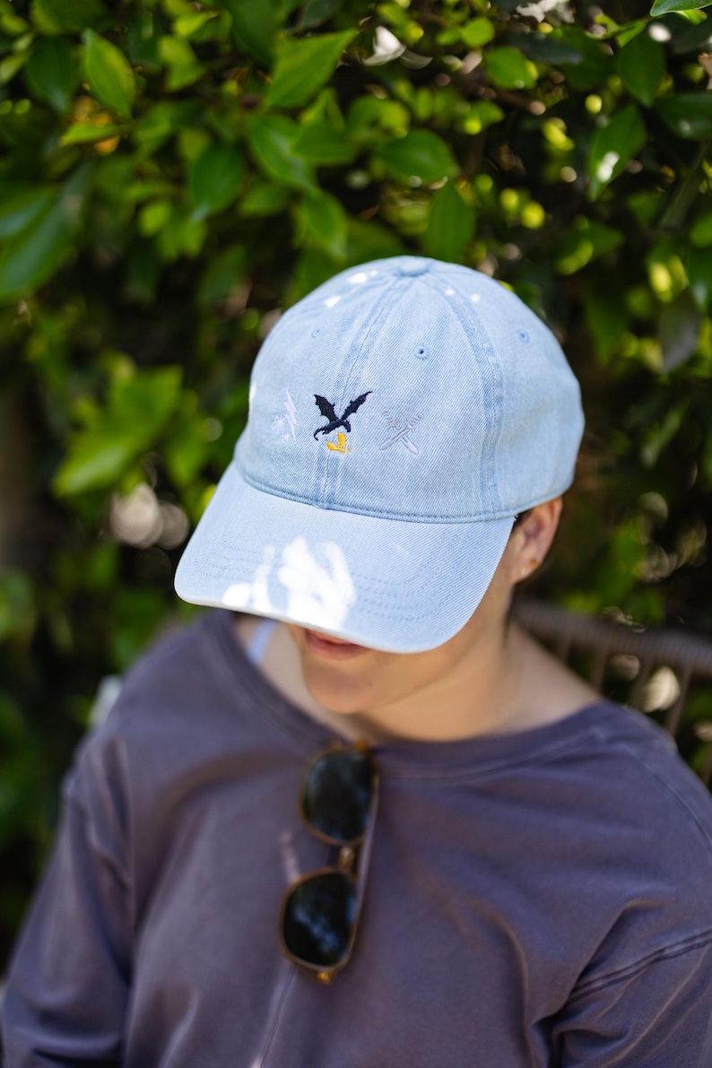 May include: A light blue denim baseball cap featuring an embroidered design on the front. The embroidery consists of a small black dragon silhouette above a yellow flame, flanked by a white lightning bolt on the left and a white sword on the right.