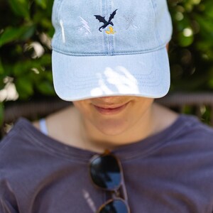 May include: A light blue denim baseball cap featuring an embroidered design on the front. The embroidery includes a white lightning bolt, a black dragon silhouette, a small yellow dragon, and two crossed silver swords in a horizontal arrangement.
