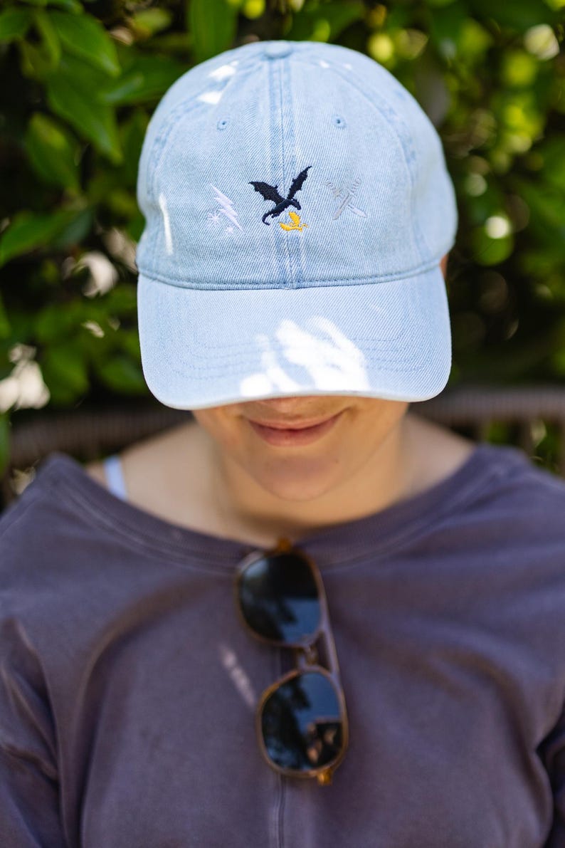 May include: A light blue denim baseball cap featuring an embroidered design on the front. The embroidery includes a white lightning bolt, a black dragon silhouette, a small yellow dragon, and two crossed silver swords in a horizontal arrangement.