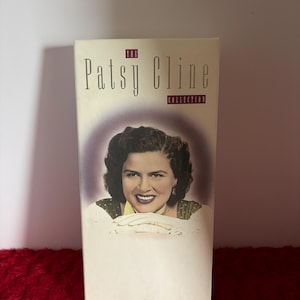 May include: A rectangular box featuring a portrait of Patsy Cline. The box has the text "The Patsy Cline Collection" in a serif font. The portrait shows Cline smiling, with dark hair and a light-colored top. The background is a soft purple.
