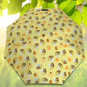 Pode incluir: Um guarda-chuva amarelo com um padrão repetido de abelhas de desenho animado, favos de mel e potes de mel. O guarda-chuva está aberto, mostrando o design. O fundo apresenta folhas verdes.