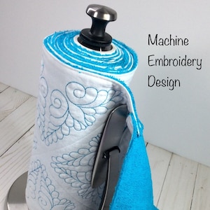 Op de afbeelding: Een witte, gestikte papieren handdoekhouder met een blauw geborduurd ontwerp. De houder is bevestigd aan een roestvrijstalen standaard met een blauwe badstof handdoek die eronder hangt. De handdoek heeft witte drukknopen op de onderste hoeken. De tekst "Machine Embroidery Design" is in zwart op een witte achtergrond geschreven. Een houten knoop met de tekst "Handmade Design" bevindt zich in de linkerbenedenhoek van de afbeelding.