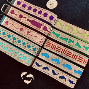 May include: Seven colorful keychains with cork backing and different designs. The designs include a wine glass, a heart, a geometric pattern, a car, a series of lines, a fish, and a clover. The keychains are all made by Mollymade Designs.