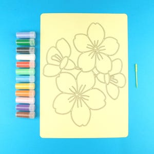 May include: A rectangular light yellow craft sheet featuring a black outline of cherry blossoms, accompanied by a vertical row of twelve small clear plastic vials filled with various coloured sands and a single thin yellow plastic applicator tool.