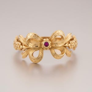 Pode incluir: Anel dourado com design de laço, detalhado com folhas e flores. Uma pequena pedra preciosa vermelha redonda está no centro do laço. O design do anel é simétrico, com detalhes florais em ambos os lados.