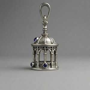 May include: A silver pendant shaped like a small gazebo, featuring a domed roof and arched openings. It has three blue gemstones and a loop for suspension. The design is highly detailed and ornate.