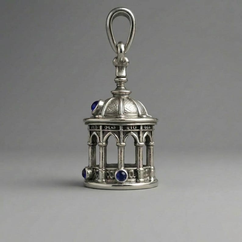 May include: A silver pendant shaped like a small gazebo, featuring a domed roof and arched openings. It has three blue gemstones and a loop for suspension. The design is highly detailed and ornate.