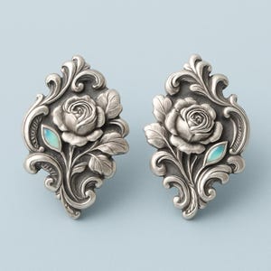 Könnte beinhalten: Ein Paar verzierte silberne Ohrringe. Jeder Ohrring zeigt ein detailliertes Rosendesign mit Blättern und verschnörkelten Verzierungen in einem dekorativen Rahmen. Ein kleiner, hellblauer Akzent ist in der Nähe der Rose gesetzt. Die Ohrringe sind vor hellblauem Hintergrund.