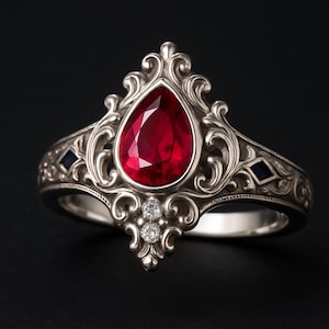 May include: A silver ring with an ornate design, featuring a large, pear-shaped red gemstone as the centerpiece. The ring is further embellished with two smaller diamonds and two dark blue square-cut stones. The band has an intricate, detailed pattern.