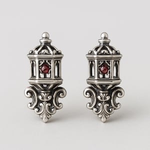 Puede incluir: Un par de pendientes plateados. Cada pendiente presenta un diseño detallado y ornamentado con una pequeña gema roja y redonda en el centro. Los pendientes tienen forma de linterna con adornos de volutas.