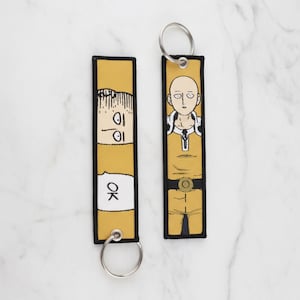 May include: Two yellow and black embroidered keychains featuring anime characters. One keychain shows a character with a bald head and yellow jumpsuit. The other keychain has a character with a worried expression and the word "OK".