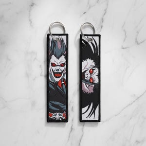 May include: Two black rectangular keychains with silver key rings. Each keychain features a colorful embroidered image of a character with red eyes and a sinister grin. One character is upright, the other is upside down.