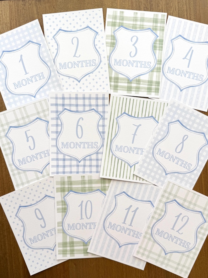 May include: Set of twelve baby milestone cards. Each card features a shield design with a number from one to twelve, representing months. The cards have various patterns, including polka dots, plaid, and stripes, in shades of blue and green.