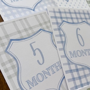 May include: A collection of baby milestone cards with a blue and white color scheme. Each card features a shield-shaped design with a number and the word "MONTHS" in a soft blue font. The cards have plaid and polka dot patterns.