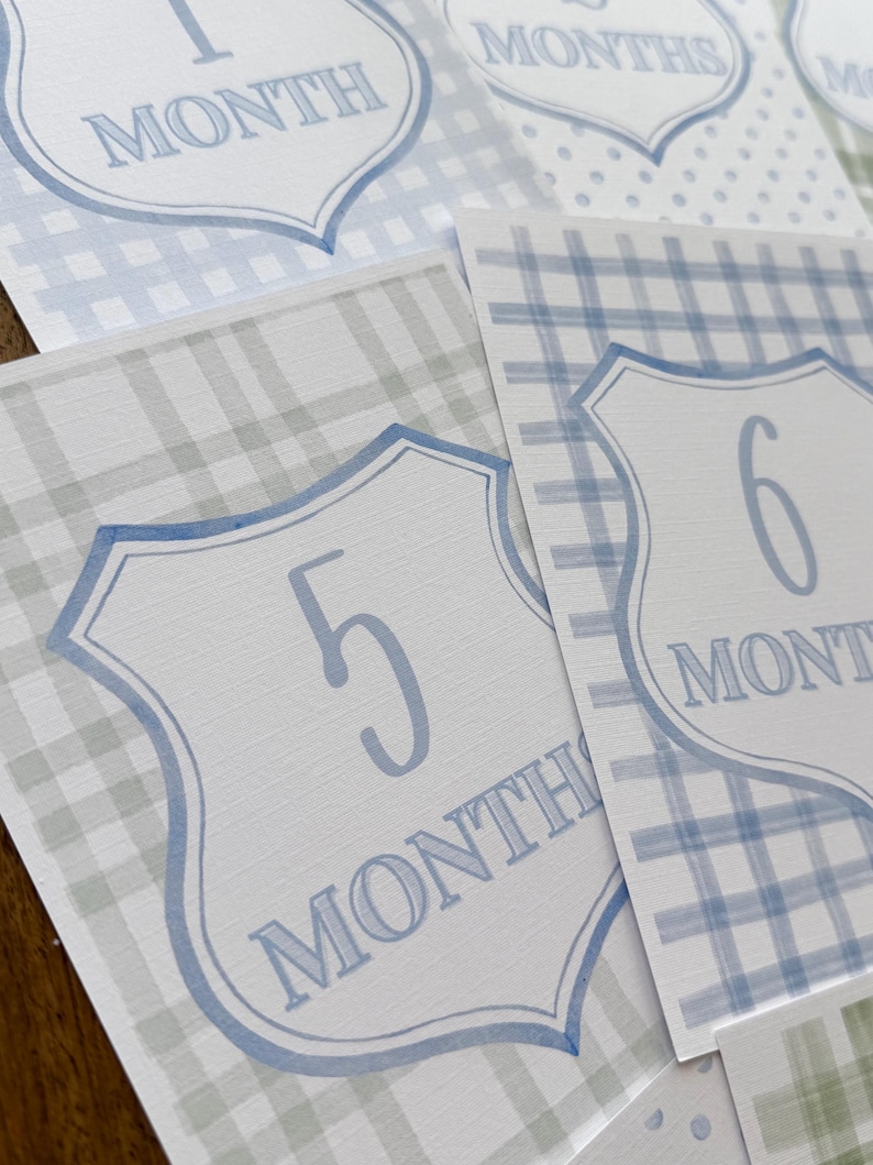 May include: A collection of baby milestone cards with a blue and white color scheme. Each card features a shield-shaped design with a number and the word "MONTHS" in a soft blue font. The cards have plaid and polka dot patterns.