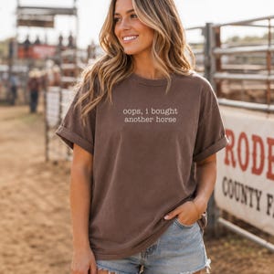 Peut inclure: Un t-shirt marron à manches courtes avec un col rond et des manches retroussées. L'avant du t-shirt présente le texte blanc "oops, i bought another horse" imprimé dans une police serif simple et minuscule au centre de la poitrine.