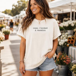 May include: An off-white, short-sleeved t-shirt featuring the black text "oops, i booked a vacation" printed on the chest. The shirt has a loose, relaxed fit with cuffed sleeves and is paired with light blue denim shorts with a frayed hem.