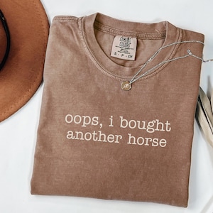 Könnte beinhalten: Ein braunes Comfort Colors T-Shirt mit dem weißen Text "oops, i bought another horse". Eine silberne Halskette mit einem kleinen Anhänger liegt über dem Shirt. Ein brauner Hut ist oben links zu sehen.