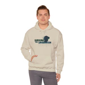 Puede incluir: Sudadera con capucha beige con bolsillo delantero y cordón. La sudadera presenta el texto "SURWING THE SIMULATION" en un diseño azul y blanco, junto con un gráfico de un casco de realidad virtual.