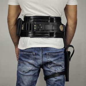 May include: Black leather utility belt with multiple pouches and a holster. The belt features gold-colored accents and is worn with blue jeans and a white t-shirt. The holster is attached to the leg with a black strap.