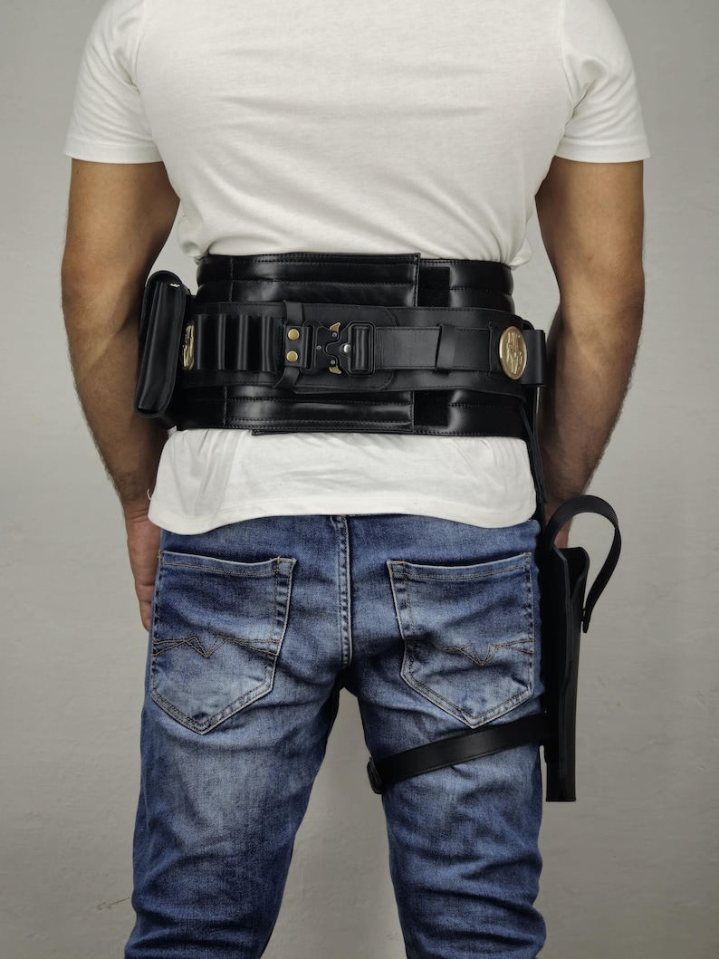 May include: Black leather utility belt with multiple pouches and a holster. The belt features gold-colored accents and is worn with blue jeans and a white t-shirt. The holster is attached to the leg with a black strap.