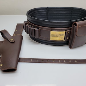 Könnte beinhalten: Ein brauner Leder-Gürtel mit Pistolenholster, Patronenhaltern und zwei Taschen. Der Gürtel hat eine goldfarbene Schnalle und eine schwarze Lederbasis. Das Holster ist für eine Handfeuerwaffe konzipiert. Der Gürtel ist zum Tragen um die Taille konzipiert.