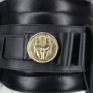 May include: Black leather belt with a gold-colored medallion featuring a stylized helmet design. The belt has a wide, layered construction with stitched detailing and a series of small loops. The medallion is circular and prominently displayed.