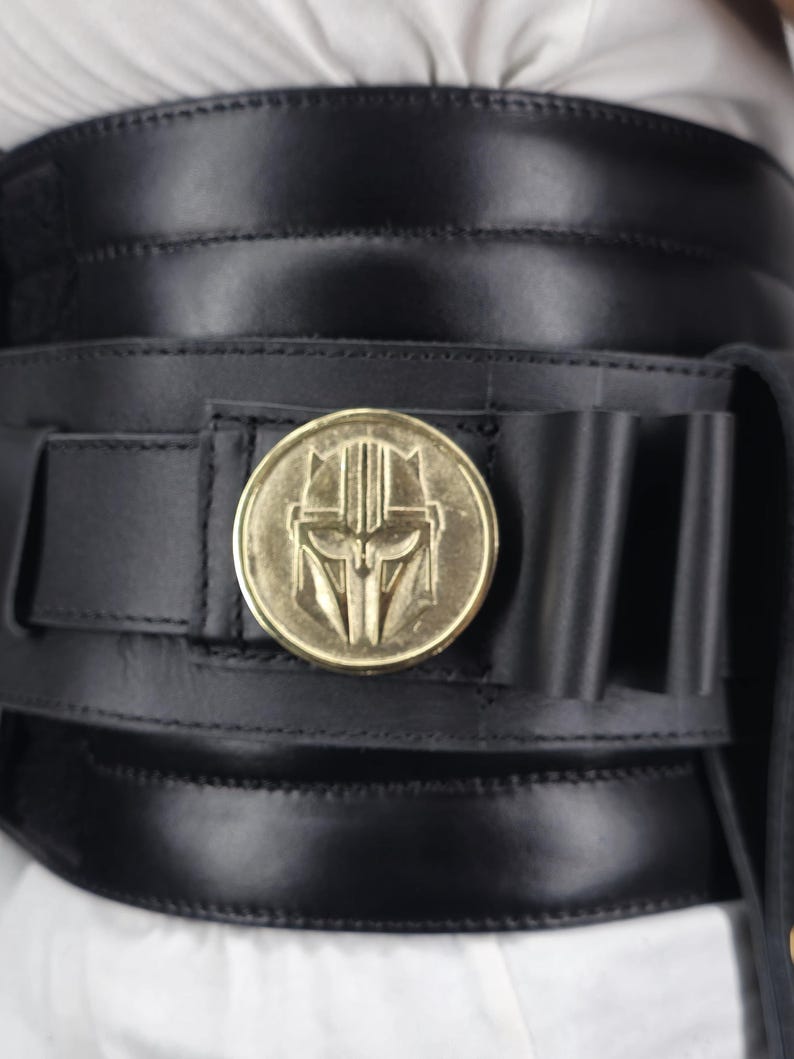 May include: Black leather belt with a gold-colored medallion featuring a stylized helmet design. The belt has a wide, layered construction with stitched detailing and a series of small loops. The medallion is circular and prominently displayed.