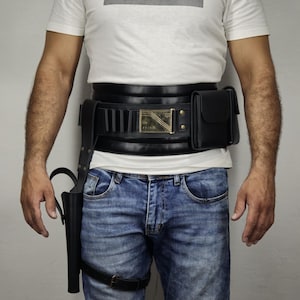 May include: A white t-shirt with the word "IMPOSSIBLE" in a black rectangle. The person is wearing a black leather belt with a holster and a pouch, and blue jeans.