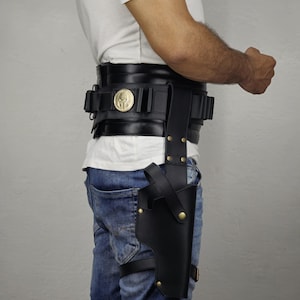 May include: Black leather gun holster attached to a belt and leg strap. The belt features a gold-colored emblem and multiple pouches. The person is wearing blue jeans and a white t-shirt.
