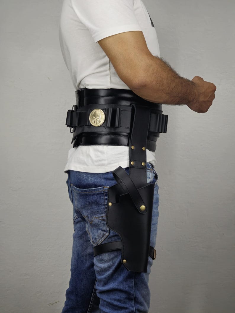 May include: Black leather gun holster attached to a belt and leg strap. The belt features a gold-colored emblem and multiple pouches. The person is wearing blue jeans and a white t-shirt.