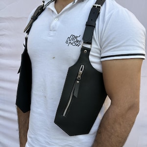 May include: Black leather shoulder holster with a silver zipper closure. The holster has adjustable straps and is worn over a white polo shirt. The shirt has the text "THE FUTURE IS NOW" printed on it.