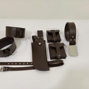 K&ouml;nnte beinhalten: Eine Sammlung brauner Lederaccessoires, darunter ein G&uuml;rtel mit silberner Schnalle, zwei Beutel, eine Messerscheide und zwei Handgelenkmanschetten. Die Artikel sind auf einer wei&szlig;en Oberfl&auml;che angeordnet.