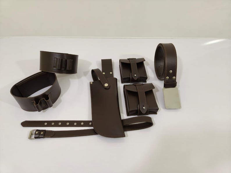 K&ouml;nnte beinhalten: Eine Sammlung brauner Lederaccessoires, darunter ein G&uuml;rtel mit silberner Schnalle, zwei Beutel, eine Messerscheide und zwei Handgelenkmanschetten. Die Artikel sind auf einer wei&szlig;en Oberfl&auml;che angeordnet.