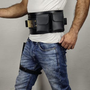 May include: Black leather utility belt with two pouches and a gold-colored buckle. The belt is worn with blue jeans and a white t-shirt. A black leather thigh strap is also visible.
