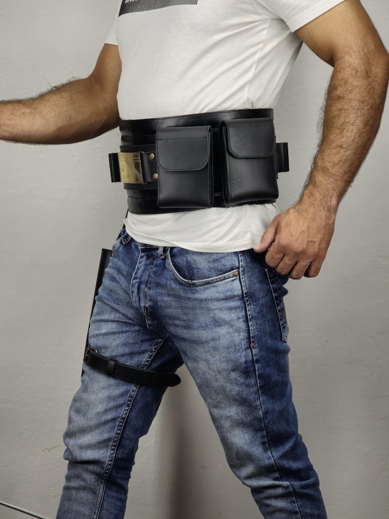 May include: Black leather utility belt with two pouches and a gold-colored buckle. The belt is worn with blue jeans and a white t-shirt. A black leather thigh strap is also visible.