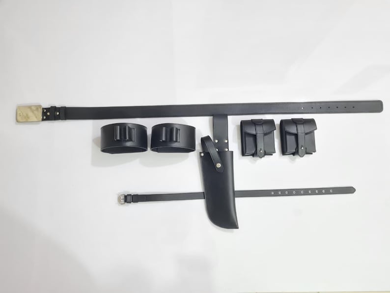 K&ouml;nnte beinhalten: Ein schwarzes Lederg&uuml;rtel-Set mit rechteckiger Schnalle, zwei Patronenhaltern, zwei kleinen Taschen, einer Messerscheide und einem kleineren G&uuml;rtel. Das Set ist f&uuml;r den Gebrauch bestimmt und liegt auf wei&szlig;em Untergrund.