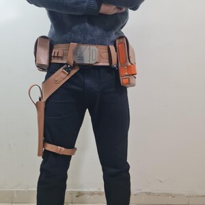 May include: A tan leather utility belt with multiple pouches and holsters, featuring a silver buckle. The belt is worn with black trousers and a grey jumper. A holster is attached to the leg.
